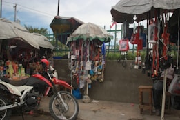 A busy street market stall with a variety of items hanging from the canopy-covered stands. A red and white motorcycle is parked in front of the stall, surrounded by various goods such as electronics, phone accessories, and personal care products. The stall is covered with makeshift umbrellas providing shade.