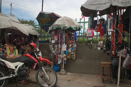 A busy street market stall with a variety of items hanging from the canopy-covered stands. A red and white motorcycle is parked in front of the stall, surrounded by various goods such as electronics, phone accessories, and personal care products. The stall is covered with makeshift umbrellas providing shade.