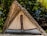 A rustic triangular hut made from natural materials with a thatched roof. The doorway is covered with a fabric curtain, while the surrounding environment includes lush green foliage and tropical trees.