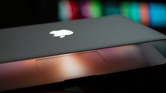 A partially closed laptop displaying an illuminated logo on the lid, with a blurred multicolored background. The trackpad area is visible with a warm glow reflecting off its surface.