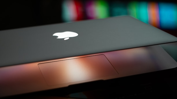 A partially closed laptop displaying an illuminated logo on the lid, with a blurred multicolored background. The trackpad area is visible with a warm glow reflecting off its surface.