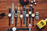 A collection of various wristwatches is arranged neatly on a wooden surface, displaying a range of designs and colors. Dice, sunglasses, a red toy car, and a yellow bottle labeled 'English Blazer Victory' are also present, adding to the display's eclectic mix.