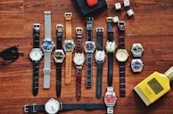 A collection of various wristwatches is arranged neatly on a wooden surface, displaying a range of designs and colors. Dice, sunglasses, a red toy car, and a yellow bottle labeled 'English Blazer Victory' are also present, adding to the display's eclectic mix.
