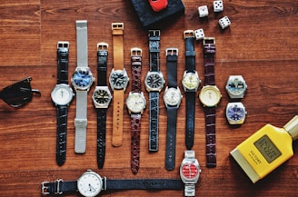 A collection of various wristwatches is arranged neatly on a wooden surface, displaying a range of designs and colors. Dice, sunglasses, a red toy car, and a yellow bottle labeled 'English Blazer Victory' are also present, adding to the display's eclectic mix.