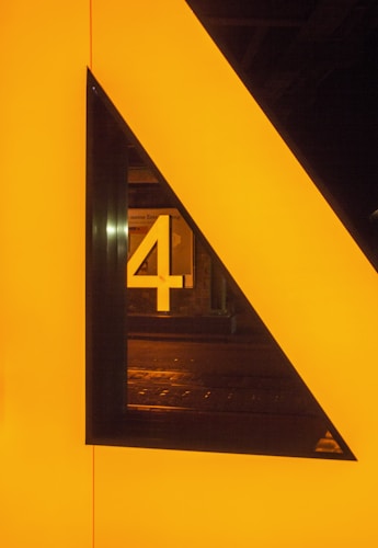 A bold, geometric design featuring a large, illuminated triangle with a bright yellow-orange color. Inside the triangle, the number 4 is prominently displayed, also lit up. The background appears to be a dark urban setting.