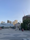 An urban scene featuring modern architecture with geometric shapes, surrounded by tall buildings. Construction cranes are visible on a high-rise, indicating ongoing development. Trees and planters add a touch of greenery to the paved plaza.