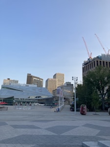 An urban scene featuring modern architecture with geometric shapes, surrounded by tall buildings. Construction cranes are visible on a high-rise, indicating ongoing development. Trees and planters add a touch of greenery to the paved plaza.