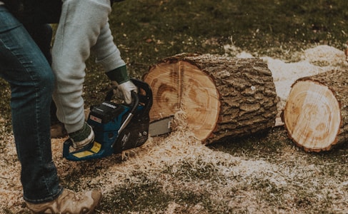 A person is using a chainsaw to cut through logs that are placed on a grassy surface. Sawdust is scattered around the freshly cut pieces of wood. The person is wearing jeans, a light-colored jacket, and sturdy boots.