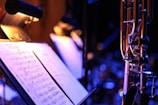 A close-up view of a music stand illuminated by a small lamp. The stand holds sheet music, which is clearly visible and legible, displaying musical notes and notations. To the right, part of a brass wind instrument is in focus, with the rest of the background slightly blurred, featuring additional stands and lights.