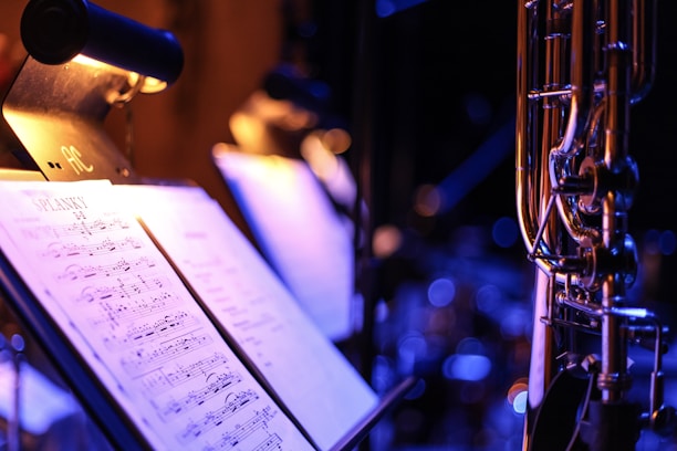 A close-up view of a music stand illuminated by a small lamp. The stand holds sheet music, which is clearly visible and legible, displaying musical notes and notations. To the right, part of a brass wind instrument is in focus, with the rest of the background slightly blurred, featuring additional stands and lights.