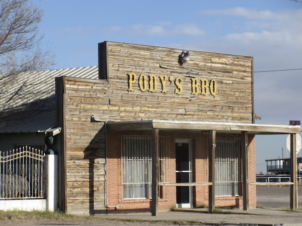 A rustic barbecue restaurant with a wooden facade displaying the sign 'Pody's BBQ'. The building has a covered entrance with metal bars on the windows and a combination of brick and wood materials. It's situated in a seemingly rural or small-town setting with clear skies.
