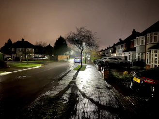 A calm residential street at night illuminated softly by streetlights, with a visible security camera in the foreground.