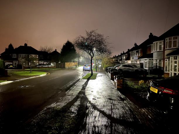A calm residential street at night illuminated softly by streetlights, with a visible security camera in the foreground.