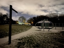 A dark and snowy playground scene with a mysterious atmosphere. The foreground features a grassy area dusted with snow, leading to a futuristic-looking metal structure with a dome-like shape. Trees line the background silhouetted against the cloudy night sky, and a row of houses lit by warm streetlights is visible in the distance.