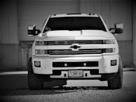 A large pickup truck parked in front of a building, featuring a prominent front grille and large tires. The truck is positioned on gravel in a monochrome setting.