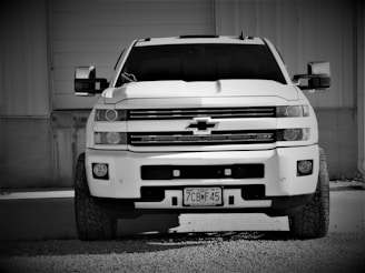 A large pickup truck parked in front of a building, featuring a prominent front grille and large tires. The truck is positioned on gravel in a monochrome setting.