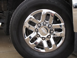 A close-up of a shiny, silver six-spoke truck wheel with the Chevrolet logo at the center, surrounded by black tire rubber engraved with the brand name Michelin. The reflective surface of the wheel captures parts of the surrounding environment.