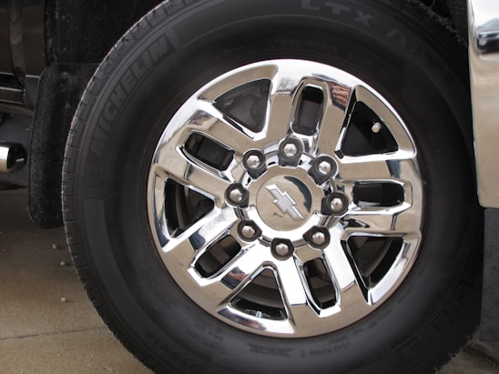 A close-up of a shiny, silver six-spoke truck wheel with the Chevrolet logo at the center, surrounded by black tire rubber engraved with the brand name Michelin. The reflective surface of the wheel captures parts of the surrounding environment.