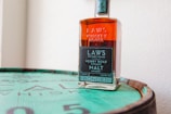 A bottle of Laws Whiskey House, labeled as Henry Road Straight Malt, is positioned on top of a whiskey barrel. The label displays information about the whiskey, including that it is bonded and aged 4 years. The background is a plain white wall, creating a focus on the bottle and its branding.