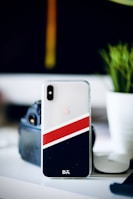 A smartphone with a sleek, transparent case displaying a diagonal design featuring red and black stripes. The device is positioned upright on a white surface. In the background, a blurred green plant and an object resembling a camera add depth to the scene.