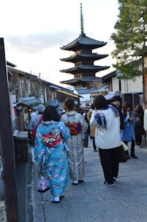 A street scene featuring people walking towards a traditional Japanese pagoda. Two women wearing colorful kimonos can be seen in the foreground, one in a blue kimono with floral patterns and the other in a light-colored kimono. The street is lined with traditional Japanese buildings, and several other people are visible, some taking photos or talking. A tall tree stands on the right side.
