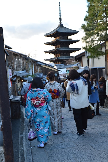 A street scene featuring people walking towards a traditional Japanese pagoda. Two women wearing colorful kimonos can be seen in the foreground, one in a blue kimono with floral patterns and the other in a light-colored kimono. The street is lined with traditional Japanese buildings, and several other people are visible, some taking photos or talking. A tall tree stands on the right side.