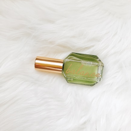 A glass bottle with a green liquid inside, labeled 'balance,' features a gold-colored cap. It is placed on a textured white fur surface.