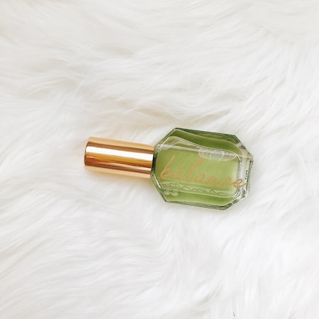 A glass bottle with a green liquid inside, labeled 'balance,' features a gold-colored cap. It is placed on a textured white fur surface.