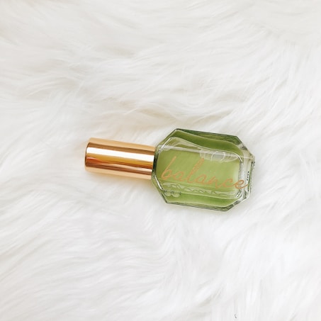 A glass bottle with a green liquid inside, labeled 'balance,' features a gold-colored cap. It is placed on a textured white fur surface.