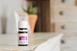 A small bottle of lavender essential oil sits on a wooden surface. The bottle has a white cap and a purple label indicating it is a therapeutic-grade product. In the blurred background, there is a plant in a terracotta pot and a light-colored wooden cabinet with drawers.