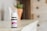 A small bottle of lavender essential oil sits on a wooden surface. The bottle has a white cap and a purple label indicating it is a therapeutic-grade product. In the blurred background, there is a plant in a terracotta pot and a light-colored wooden cabinet with drawers.