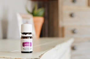 A small bottle of lavender essential oil sits on a wooden surface. The bottle has a white cap and a purple label indicating it is a therapeutic-grade product. In the blurred background, there is a plant in a terracotta pot and a light-colored wooden cabinet with drawers.