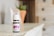 A small bottle of lavender essential oil sits on a wooden surface. The bottle has a white cap and a purple label indicating it is a therapeutic-grade product. In the blurred background, there is a plant in a terracotta pot and a light-colored wooden cabinet with drawers.