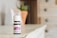 A small bottle of lavender essential oil sits on a wooden surface. The bottle has a white cap and a purple label indicating it is a therapeutic-grade product. In the blurred background, there is a plant in a terracotta pot and a light-colored wooden cabinet with drawers.