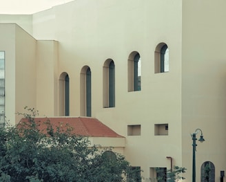 A building facade with several tall, arched windows and a modern architectural style. The structure features a flat, light-colored surface contrasted with a red-tiled roof. Greenery is visible in the foreground, along with a vintage-style street lamp.