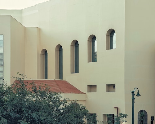 A building facade with several tall, arched windows and a modern architectural style. The structure features a flat, light-colored surface contrasted with a red-tiled roof. Greenery is visible in the foreground, along with a vintage-style street lamp.