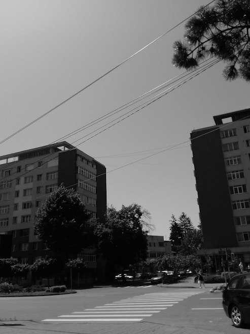 Urban scene featuring several tall residential buildings with trees lining the streets below. Power lines cross the sky, and a zebra crossing is visible in the foreground. A few cars and pedestrians are present, suggesting a typical day in a city environment.