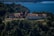 A large, rustic hotel nestled in a dense forested area overlooking a vast blue lake. The building features traditional architecture with sloped roofs and multiple chimneys, surrounded by lush greenery.