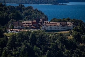 A large, rustic hotel nestled in a dense forested area overlooking a vast blue lake. The building features traditional architecture with sloped roofs and multiple chimneys, surrounded by lush greenery.