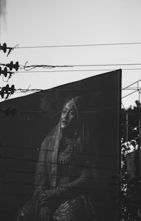 A black and white photograph captures a large billboard featuring a person in traditional attire. The billboard is partially obscured by overhead power lines crossing horizontally. The person's expression and pose suggest elegance and poise. The image is set against a clear sky with silhouettes of utility structures.