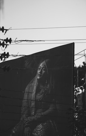 A black and white photograph captures a large billboard featuring a person in traditional attire. The billboard is partially obscured by overhead power lines crossing horizontally. The person's expression and pose suggest elegance and poise. The image is set against a clear sky with silhouettes of utility structures.