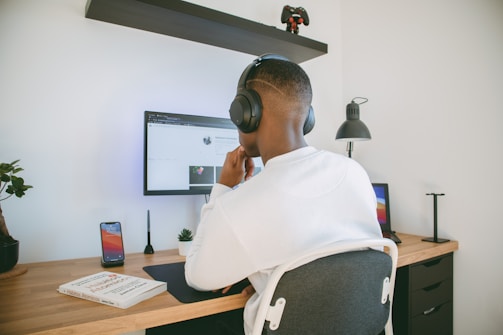 A person wearing headphones is sitting in front of a computer screen on a wooden desk. A smartphone, a lamp, and a closed book are placed on the desk. The person appears to be focused on the screen. A gaming controller is displayed above on a wall shelf.