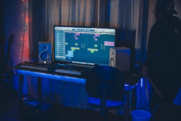 A dimly lit room contains a music production setup with a large monitor displaying a digital audio workstation interface. Surrounding the monitor are two speakers, a keyboard, and various electronic equipment. A string of colored lights adds ambiance, and a person is partially visible, appearing to be interacting with the setup.
