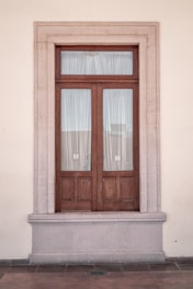A symmetrical wooden door set in a stone frame on a beige wall. The door has two panels covered with sheer white curtains.
