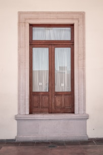 A symmetrical wooden door set in a stone frame on a beige wall. The door has two panels covered with sheer white curtains.