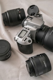 A DSLR camera with a silver and black body is placed on a light-colored fabric surface. Surrounding the camera are three camera lenses with black caps and bodies, arranged in a scattered manner. The scene conveys a sense of readiness for photography.