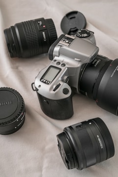A DSLR camera with a silver and black body is placed on a light-colored fabric surface. Surrounding the camera are three camera lenses with black caps and bodies, arranged in a scattered manner. The scene conveys a sense of readiness for photography.
