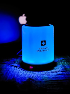 A glowing blue cylindrical object with the brand Swiss Military written on it is set against a dark background. To the left, a partially visible illuminated logo resembling an apple is present.