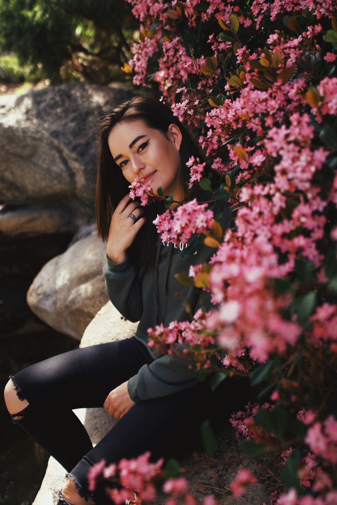 woman in black long sleeve shirt and black pants sitting on rock near pink flowers