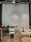 A modern cafe interior with industrial design elements featuring exposed ceilings and concrete walls. A circular metal grid dominates the wall with a neon sign at its center. Several chairs and tables are scattered around the room, with a variety of seating options including stools and regular chairs. Hanging pendant lights provide illumination.